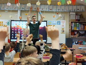 In einem Klassenraum. Ein Erwachsener steht vorne vor den Kindern an ihren Schulbänken. Alle strecken die Arme nach oben.