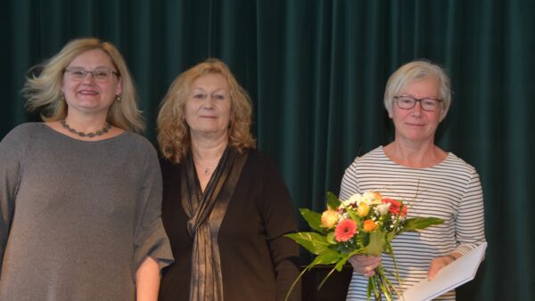 Drei Frauen vor dunkelgrünem Vorhand. Eine ältere Frau mit Blumenstrauß und Zeugnis in den Händen. Glückliche Gesichter.