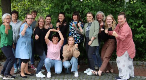 14 Frauen im Gruppenbild. Draußen vor Sträuchern. Die Frauen haben dynamische Posen eingenommen, geballte Fäuste und lächeln fröhlich und zuversichtlich.