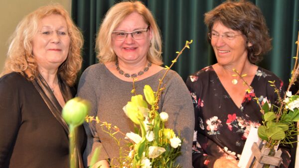 Drei Frauen vor dunkelgrünem Vorhand mit kleinen Blumengestecken in der Hand. Alle lächeln fröhlich.