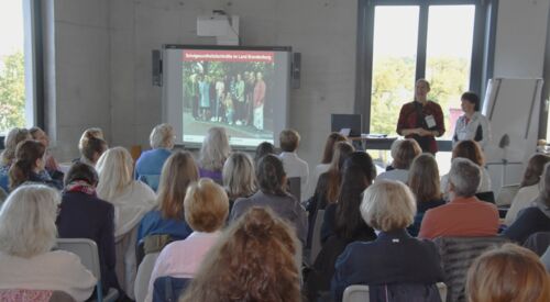 Großer Raum, viele Stuhlreihen ca. 30 Menschen sitzen im Raum. Vorn stehen zwei Frauen, eine Präsentation läuft auf einer Leinwand.