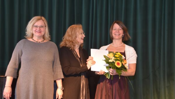 Drei Frauen vor dunkelgrünem Vorhand. Eine Frau mit brauen langen Haaren, Brille und Zeugnis in der Hand, lächelt in die Kamera. Alle lächeln und freuen sich.