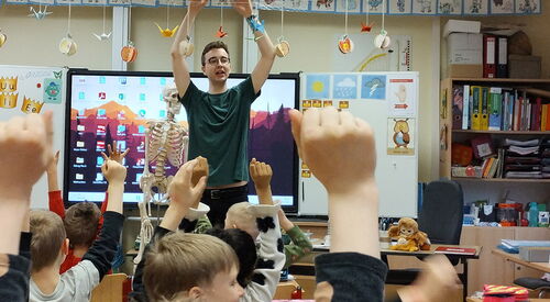 In einem Klassenraum. Ein Erwachsener steht vorne vor den Kindern an ihren Schulbänken. Alle strecken die Arme nach oben.