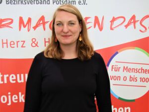 Portrait. Frau vor AWO Aufsteller. Sie hat schulterlange braune Haare, die sie offen trägt. Gelb-rote kleine Ohrringe und ein schlichtes langärmliges schwarzes Oberteil.