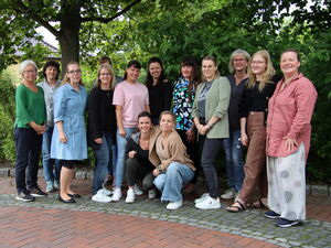 Draußen unter Bäumen. 11 Frauen stehen versetzt nebeneinander, in der Mitte sind zwei Frauen in die Hocke gegangen. Alle lächeln fröhlich in die Kamera.