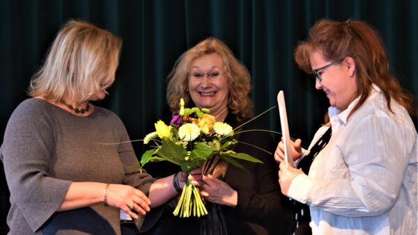 Drei Frauen vor dunkelgrünem Vorhand. Blumenstrauß und Zeugnis werden an weibliche Schulgesundheitsfachkraft übergeben. Glückliche Gesichter.